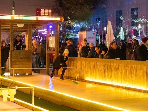 People are standing by a curling rink, shooting curling stones along the track. The area is lit up with fairy lights and it is evening.