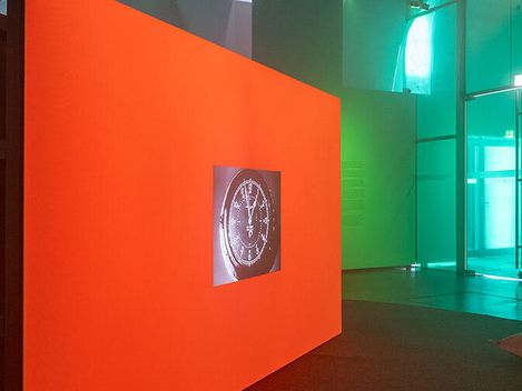 Dunkler Raum mit einer großen, leuchtend orangen Wand, auf der ein rundes Bild projiziert wird, umgeben von Kissen auf dem Boden und Glaswänden im Hintergrund.