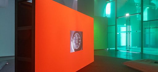 Dunkler Raum mit einer großen, leuchtend orangen Wand, auf der ein rundes Bild projiziert wird, umgeben von Kissen auf dem Boden und Glaswänden im Hintergrund.