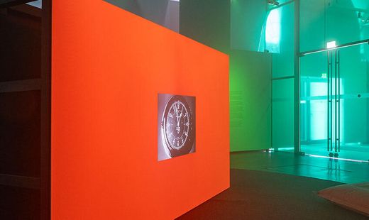 Dimly lit room with a large bright orange wall displaying a circular projected image, surrounded by cushions on the floor and glass walls in the background.