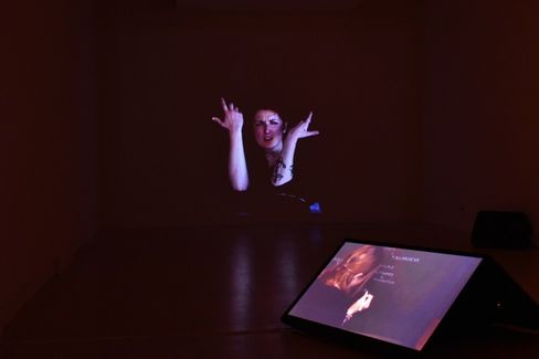In a dark room, a projection of a woman with raised hands is visible, with an illuminated screen showing a similar image in front.