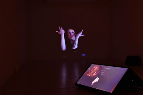 In a dark room, a projection of a woman with raised hands is visible, with an illuminated screen showing a similar image in front.