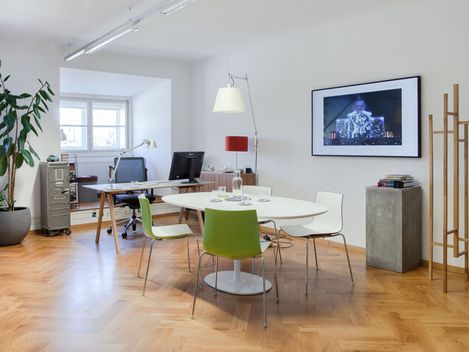 Moderner, heller Büroraum mit rundem Tisch, mehreren Stühlen, Schreibtisch mit Computer, großem Fenster, Pflanzen und minimalistischem Dekor