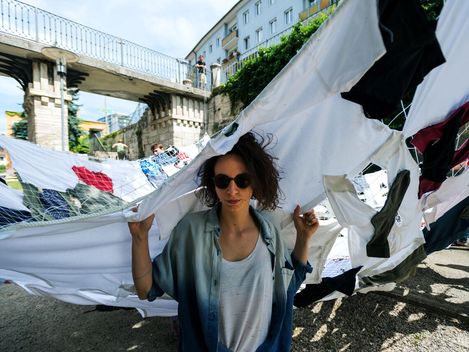 Frau mit Sonnenbrille hält ein großes, zerrissenes Tuch über sich, im Hintergrund Brücke und weitere Personen.