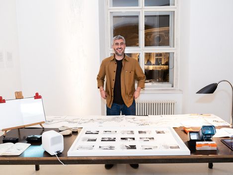Person standing at a table with spread-out drawings and prints in a bright room with two windows and a radiator.