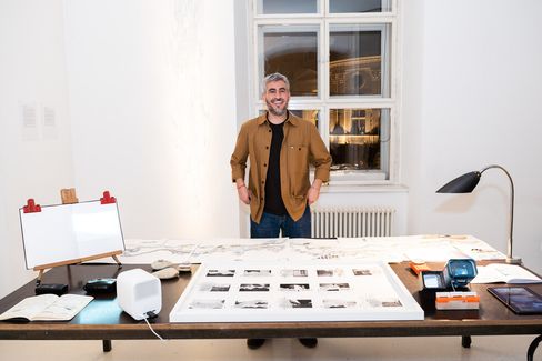 Person standing at a table with spread-out drawings and prints in a bright room with two windows and a radiator.