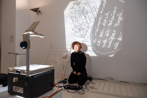 A person kneeling in front of a wall with abstract sketches and text projected by an overhead projector. The scene is artistic and creative.