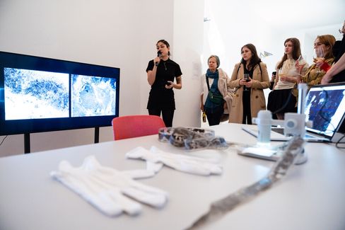 A person speaks into a microphone near a screen displaying blue and white abstract images. A group of people attentively listen, while gloves and equipment are on a nearby table. The scene conveys a thoughtful and engaged atmosphere.