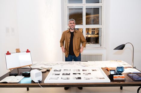 Person standing at a table with spread-out drawings and prints in a bright room with two windows and a radiator.