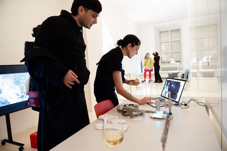 Two individuals in dark clothing interact at a table with a laptop, art tools, and a wine glass. The setting is a white, contemporary gallery space with bright lighting.