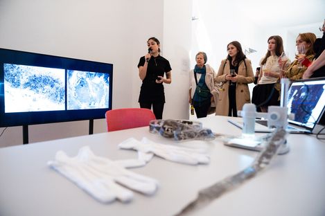A person speaks into a microphone near a screen displaying blue and white abstract images. A group of people attentively listen, while gloves and equipment are on a nearby table. The scene conveys a thoughtful and engaged atmosphere.