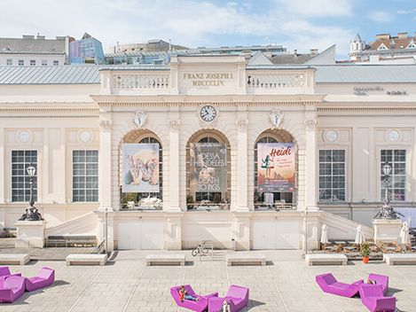 Fassade eines historischen Gebäudes und drei großen Plakaten in den Arkaden, davor mehrere violette Liegestühle auf einem gepflasterten Platz