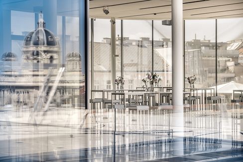 Modernes Glasgebäude mit Spiegelungen im Innenbereich, mit Blumenvasen auf Tischen. Der Blick vom Dach zeigt eine historische Kuppelkonstruktion, die eine ruhige Atmosphäre schafft.