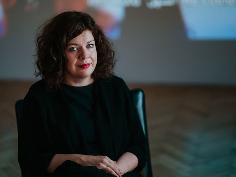 Woman with curly dark hair and red lipstick sitting in a chair against a blurred background.