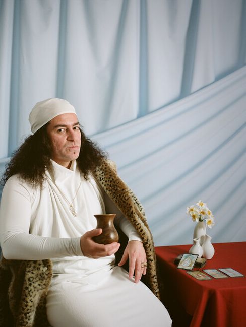 Person dressed in white with a headscarf holding a cup seated beside a red-covered table with a small vase and cards