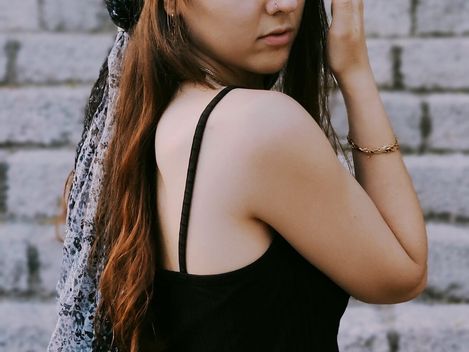 Frau mit langem Haar, schwarzem ärmellosem Oberteil, senffarbenem Rock und gemustertem Kopftuch steht seitlich vor einer Steintreppe.