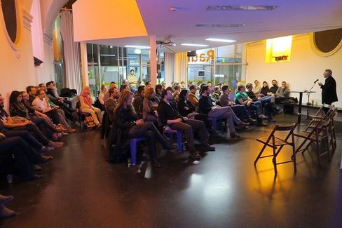 Group of people sitting in a room listening to a standing person speaking into a microphone