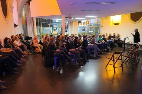 Group of people sitting in a room listening to a standing person speaking into a microphone