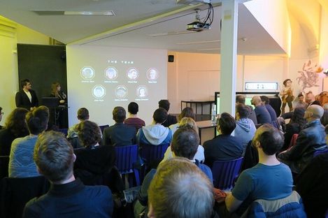 Several people sit in a room watching a wall projection showing team photos and names while two people present at the front.