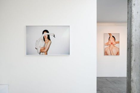 Two photographs on white gallery walls, each showing a woman with bare shoulders, the left one with a white fabric covering her face, the right one with crossed arms in front of her face.