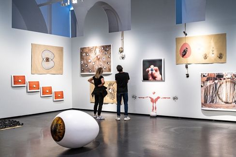 Two people stand in a gallery with high vaulted ceilings looking at various artworks on the walls and a large eye-shaped sculpture on the floor