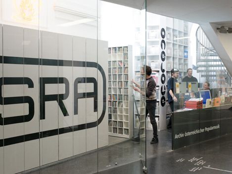 Glass-walled archive room with a large SRA logo, shelves, and people inside.