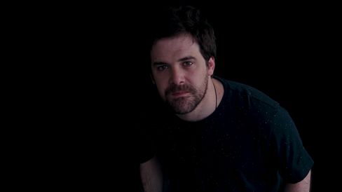 Portrait of a man with dark hair and beard against a black background.
