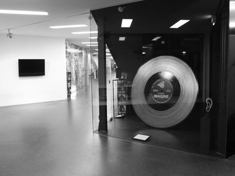 Schwarz-weiße Aufnahme eines Museumsraums mit einer großen Schallplatte hinter Glas und einem Bildschirm an der Wand