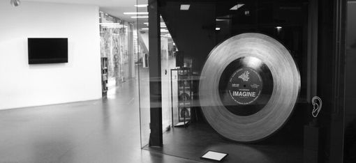 Schwarz-weiße Aufnahme eines Museumsraums mit einer großen Schallplatte hinter Glas und einem Bildschirm an der Wand