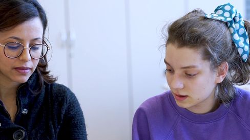 Two women sitting side by side, one wearing glasses and a dark sweater, the other with a blue hairband with white dots, both looking down.