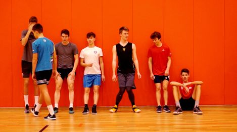 Group of seven teenagers in sportswear standing and sitting against an orange wall.
