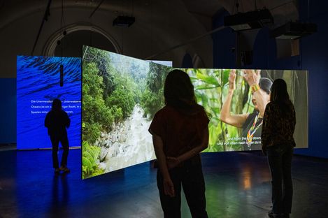 Three people stand in a dark room in front of large illuminated screens showing nature scenes and text.