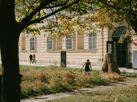 Person mit schwarzer Kleidung mäht Gras vor einem hellen Gebäude mit großen Fenstern und einem Eingang mit Schild