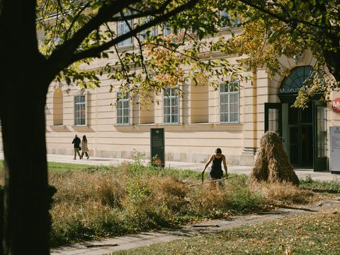Person mit schwarzer Kleidung mäht Gras vor einem hellen Gebäude mit großen Fenstern und einem Eingang mit Schild