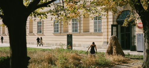 Person mit schwarzer Kleidung mäht Gras vor einem hellen Gebäude mit großen Fenstern und einem Eingang mit Schild