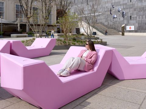 A woman in a rose-colored blazer sits relaxed on pink geometric seating called Enzis in the main courtyard of the MuseumsQuartier
