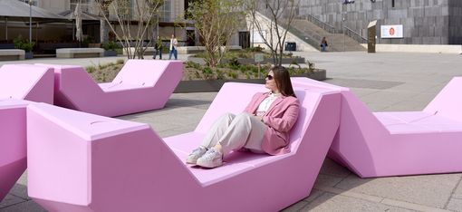 A woman in a rose-colored blazer sits relaxed on pink geometric seating called Enzis in the main courtyard of the MuseumsQuartier