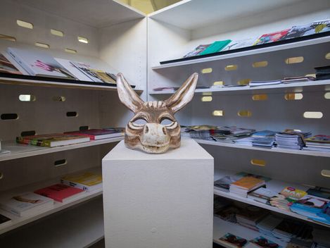 Sculpture of a donkeyhead on a white pedestal in a room with shelves filled with magazines