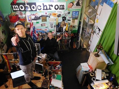 Two people in a cluttered room filled with posters, books, and technical equipment, one person pointing forward with an outstretched arm