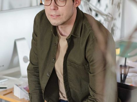 Junger Mann mit zerzaustem Haar und Brille sitzt in einem Arbeitszimmer mit Schreibtisch und Regalen im Hintergrund.
