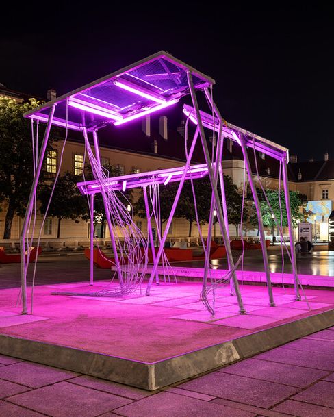 Solarpanel aus dünnen Metallstangen mit zwei horizontalen, von pinkem Licht beleuchteten Ebenen auf einem quadratischen Podest in einem nächtlichen urbanen Umfeld