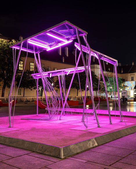 Solarpanel aus dünnen Metallstangen mit zwei horizontalen, von pinkem Licht beleuchteten Ebenen auf einem quadratischen Podest in einem nächtlichen urbanen Umfeld