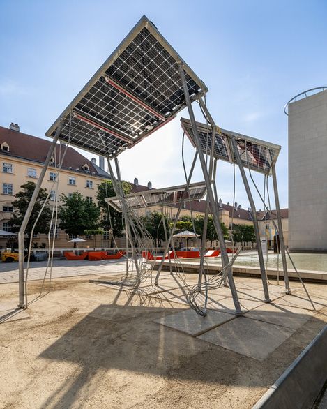 Drei große Solarpaneele auf schrägen Metallgestellen in einem städtischen Platz mit Gebäuden im Hintergrund