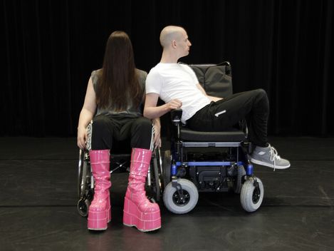 Zwei Personen in Rollstühlen sitzen Rücken an Rücken auf einer dunklen Bühne, eine trägt auffällige hohe Plateau-Stiefel.