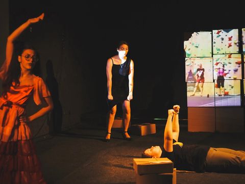 Three people on a stage, one standing with raised arm wearing a dress, another standing in a black dress, the third lying on the floor with raised legs, a screen with colorful images in the background.