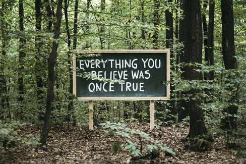 A sign with the text "EVERYTHING YOU BELIEVE WAS ONCE TRUE" stands in a forest among trees and foliage.