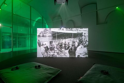Schwarz-weiße Projektion eines historischen Fotos mit Menschen auf Booten in einem dunklen Raum mit grüner Beleuchtung und Sitzkissen auf dem Boden
