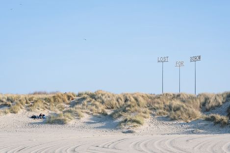 Sand dunes with grass and three tall poles displaying the words "LOST", "FOR", and "2030W".