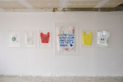 Six transparent plastic bags containing various colored and inscribed objects hang on a white wall in a room with visible ceiling structure.