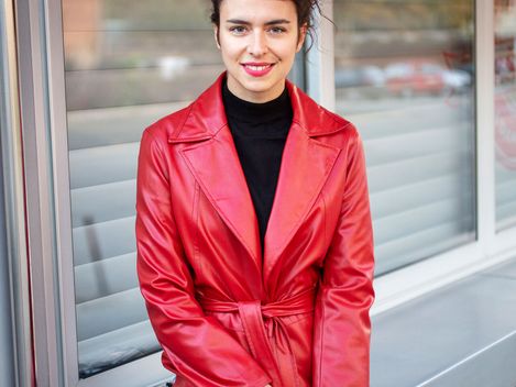 Woman with dark hair wearing a red coat sitting in front of a window with closed blinds smiling at the camera.